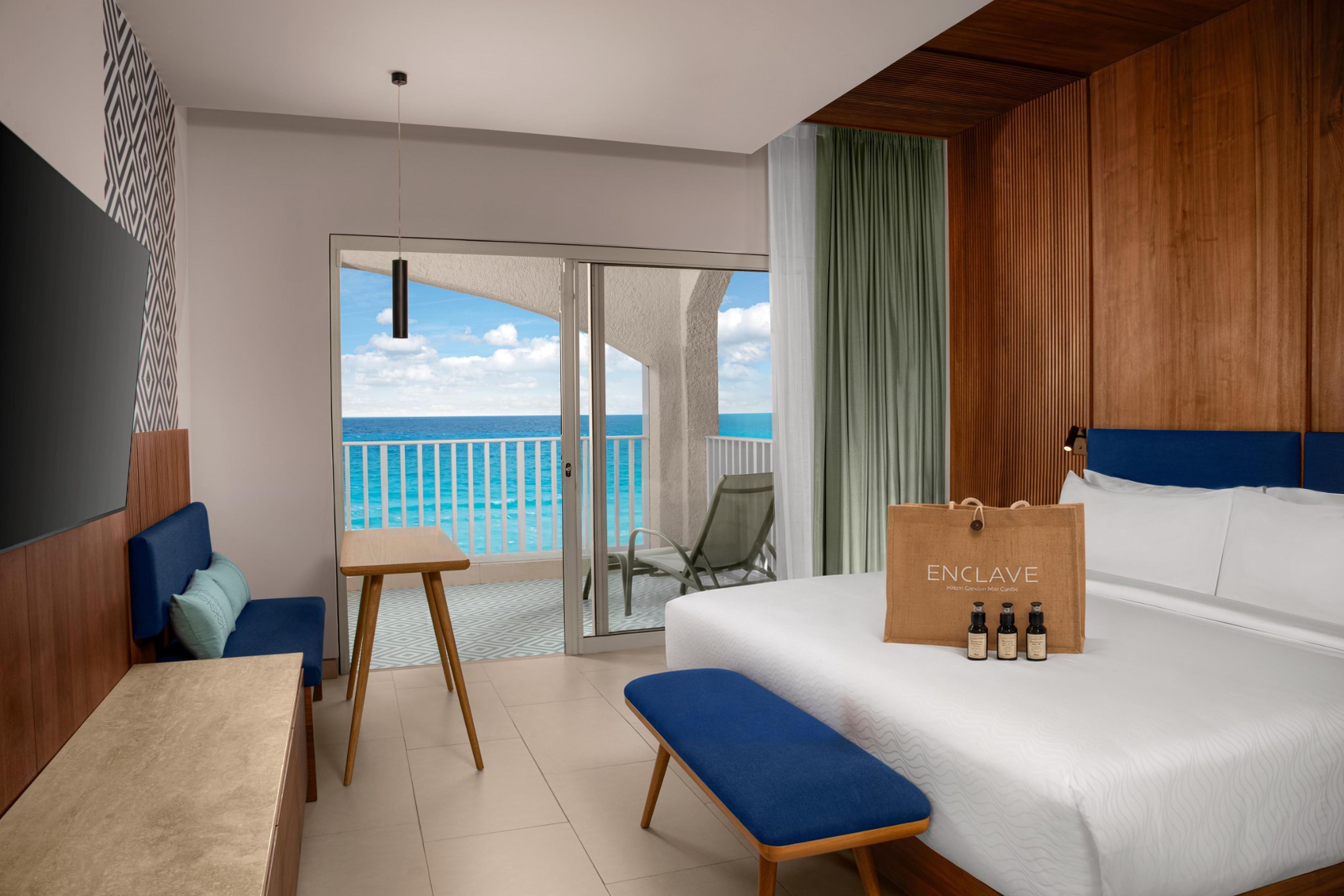 Ett hotellrum med en säng, en bänk, ett bord, en stol och utsikt över havet genom en skjutdörr.