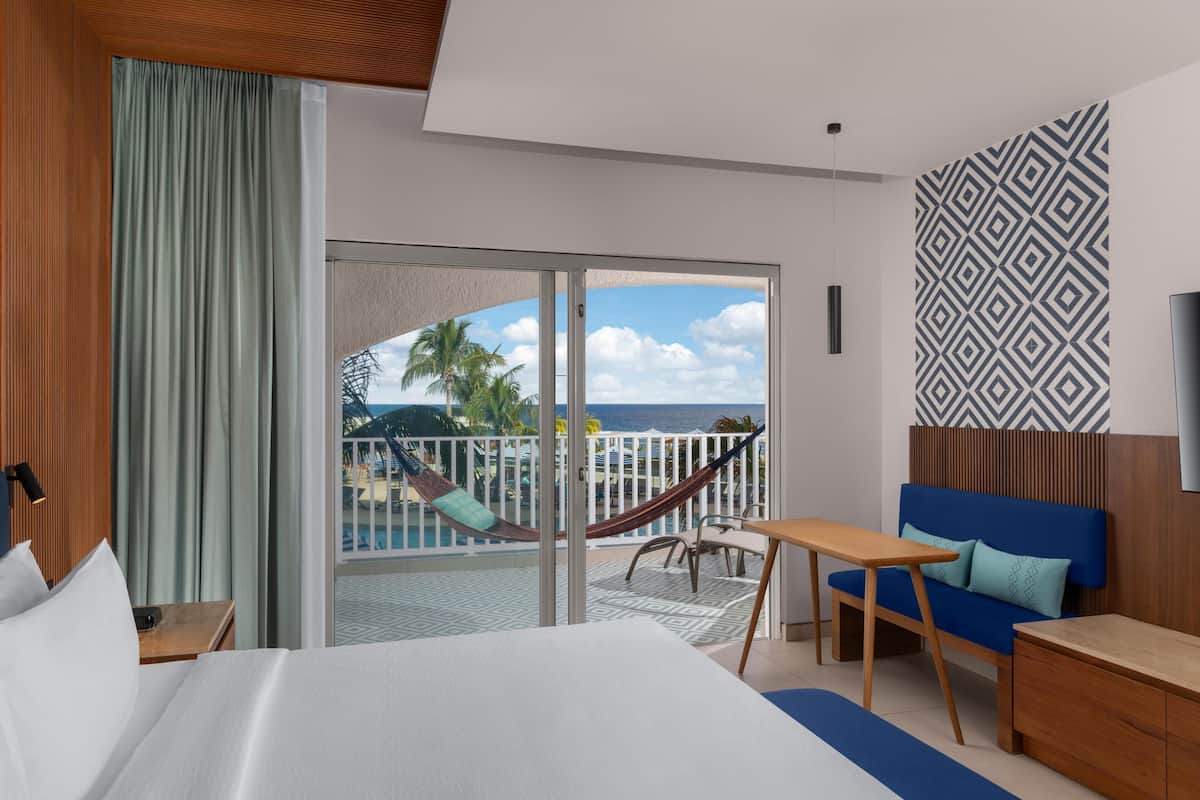 A hotel room with a bed, a sofa, a table, and a balcony with a view of the ocean and palm trees.