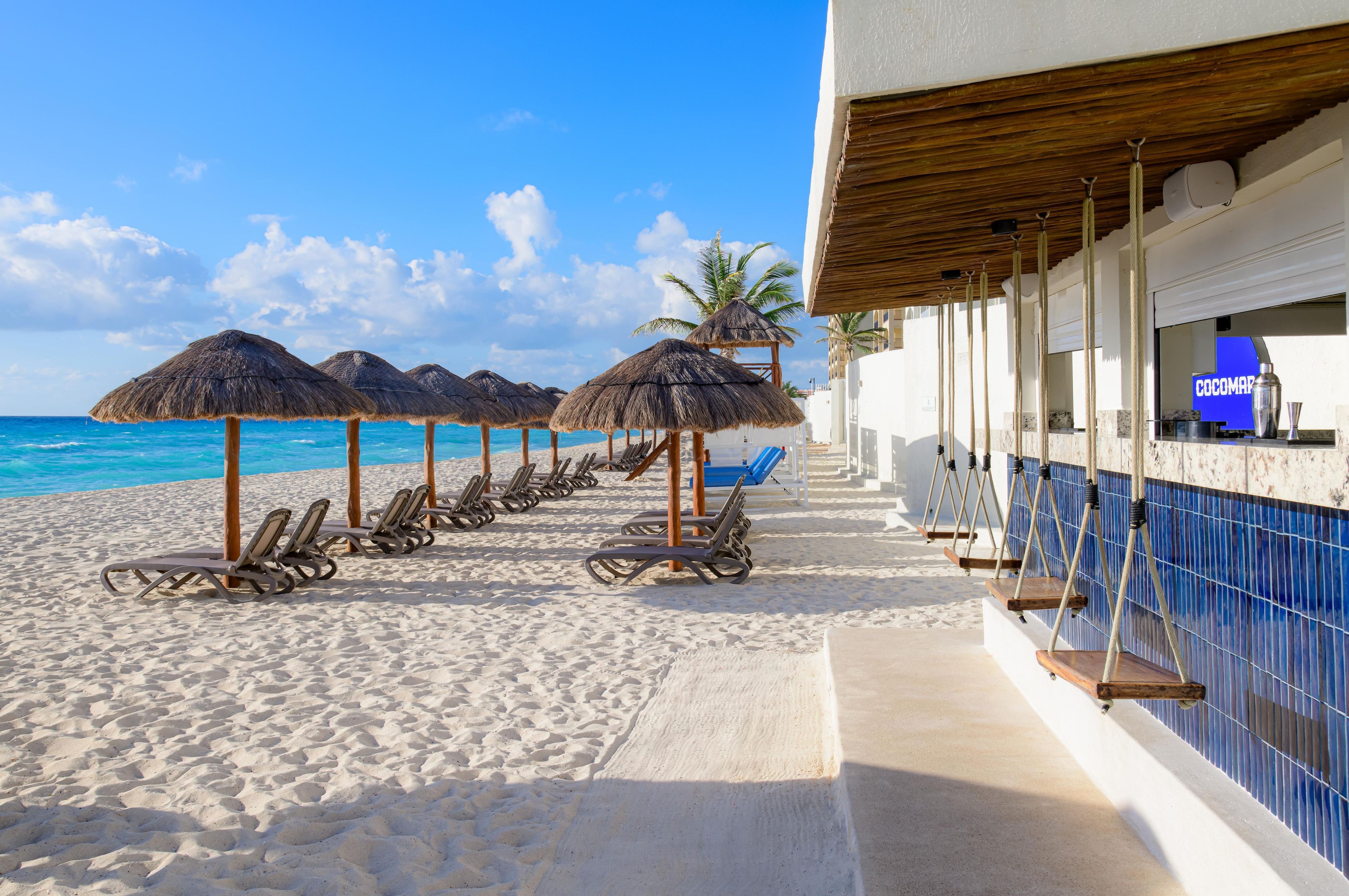 Un hotel frente a la playa con tumbonas, sombrillas de paja y una zona de bar con vendedor de cocadas.