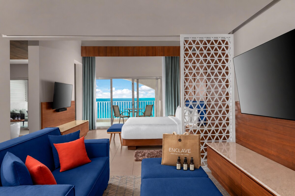 A hotel room with a bed, a sofa, a TV, a balcony with a view of the sea, and a luggage bag on the floor.