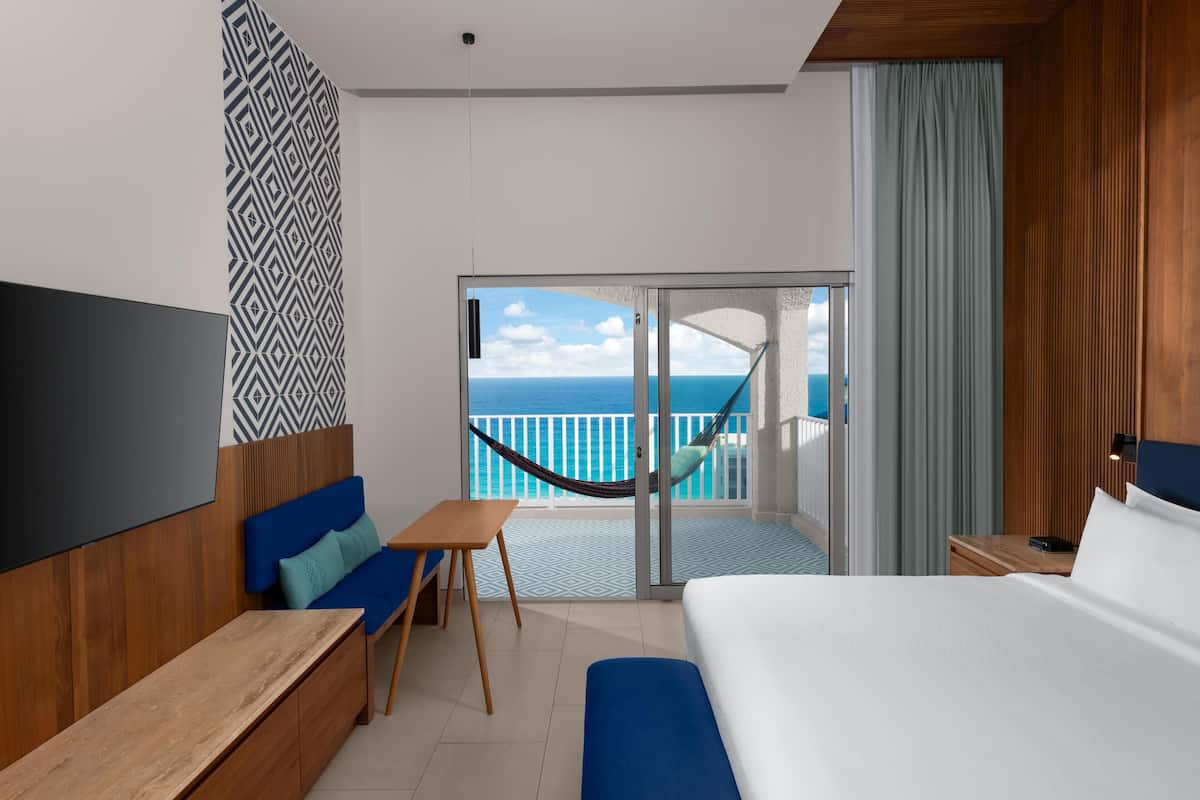 A hotel room with a bed, a desk, a chair, a TV, a balcony with a view of the sea, and a patterned wall.