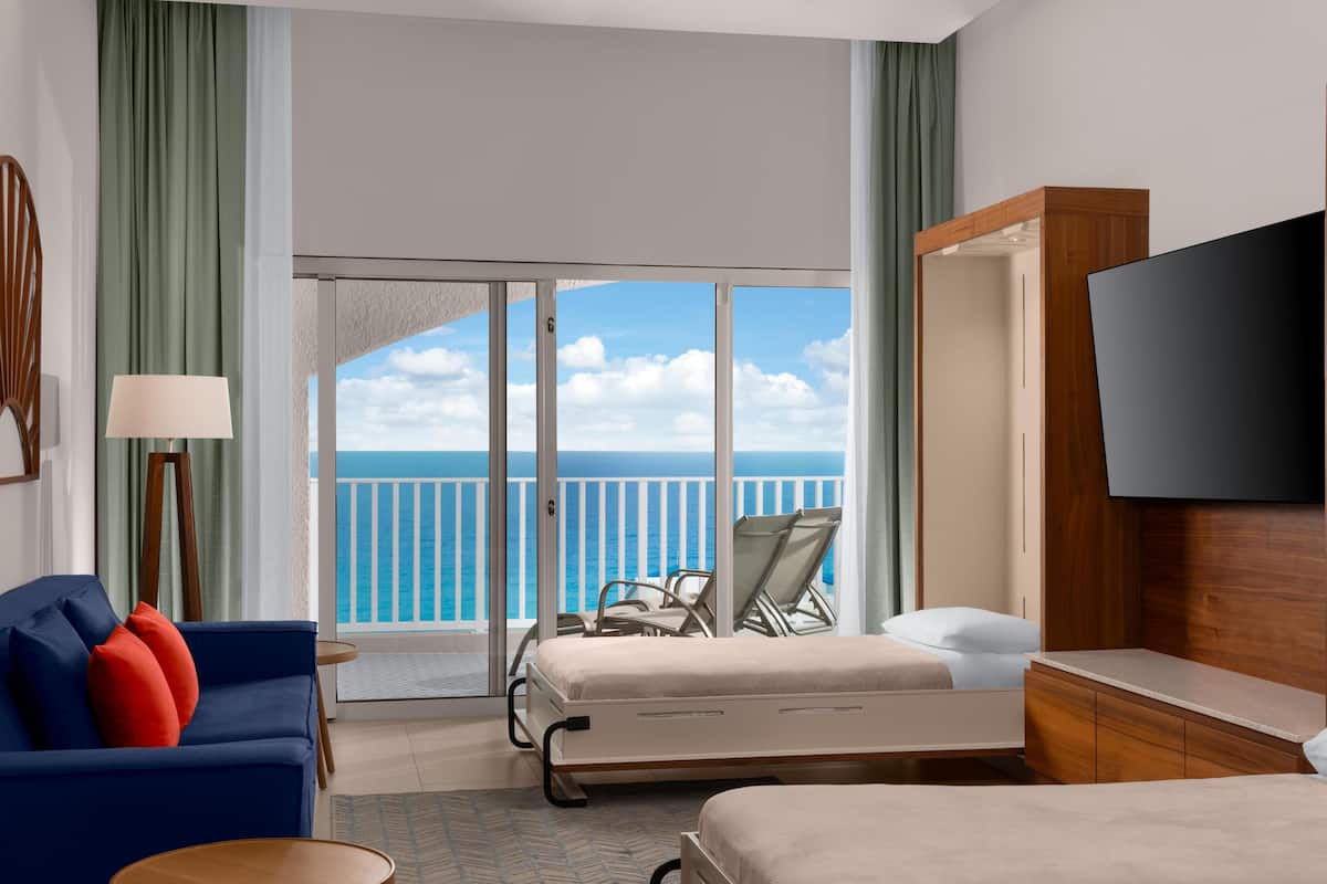 A hotel room with a bed, a sofa with red cushions, a TV, a balcony with a view of the ocean, and a wooden desk.
