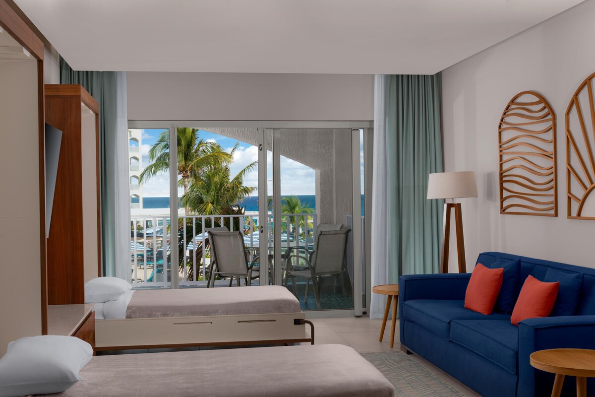 A hotel room with a bed, a blue sofa, a small table, and a view of the ocean and palm trees through a sliding glass door.