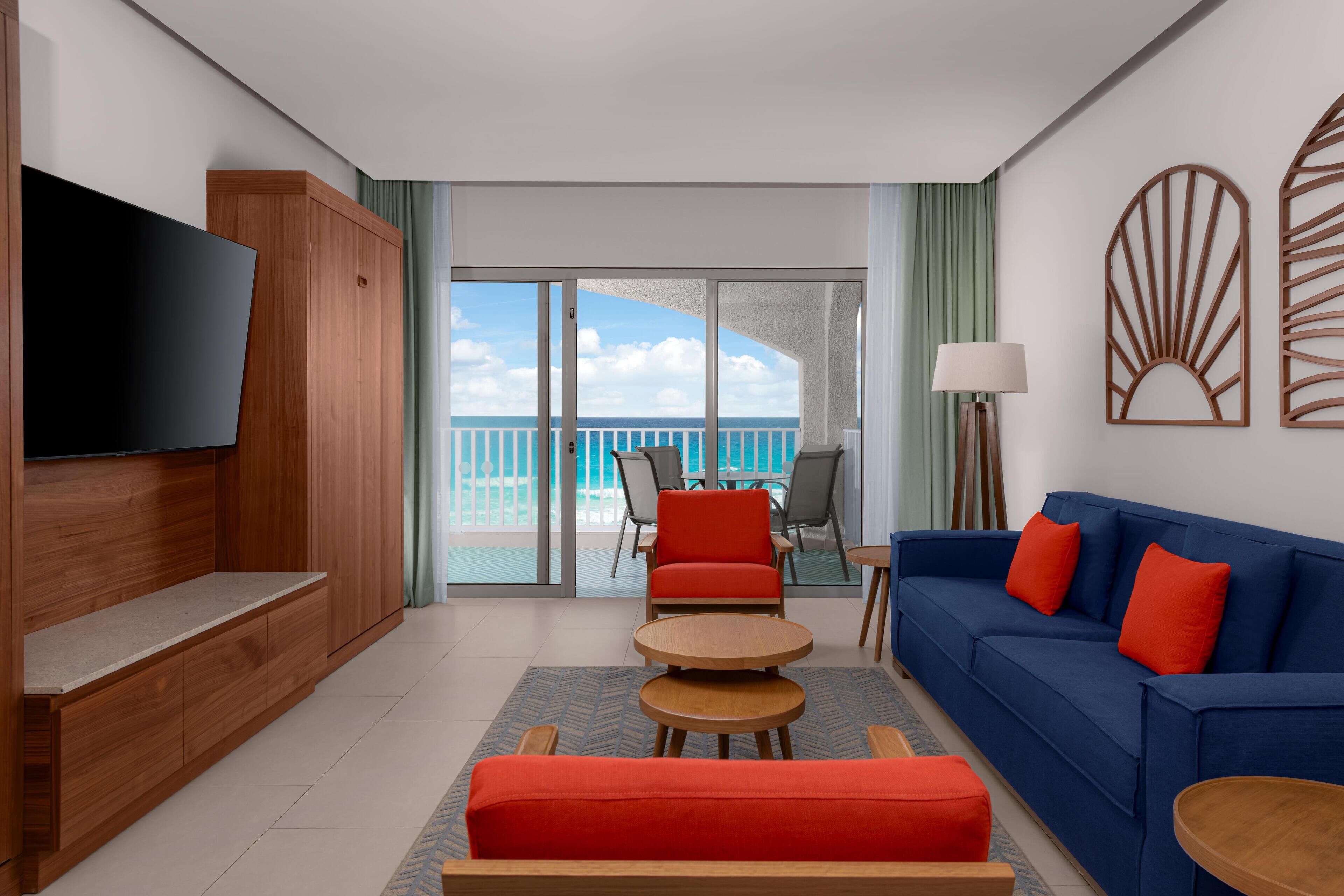 Una sala de estar moderna con televisor, muebles de madera, un sofá azul con cojines rojos y vista al mar a través de puertas de vidrio grandes.