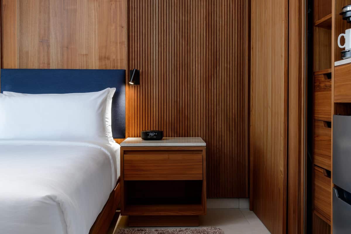 A neatly made bed with white linens, a wooden nightstand with a digital clock, and a wooden panel wall.