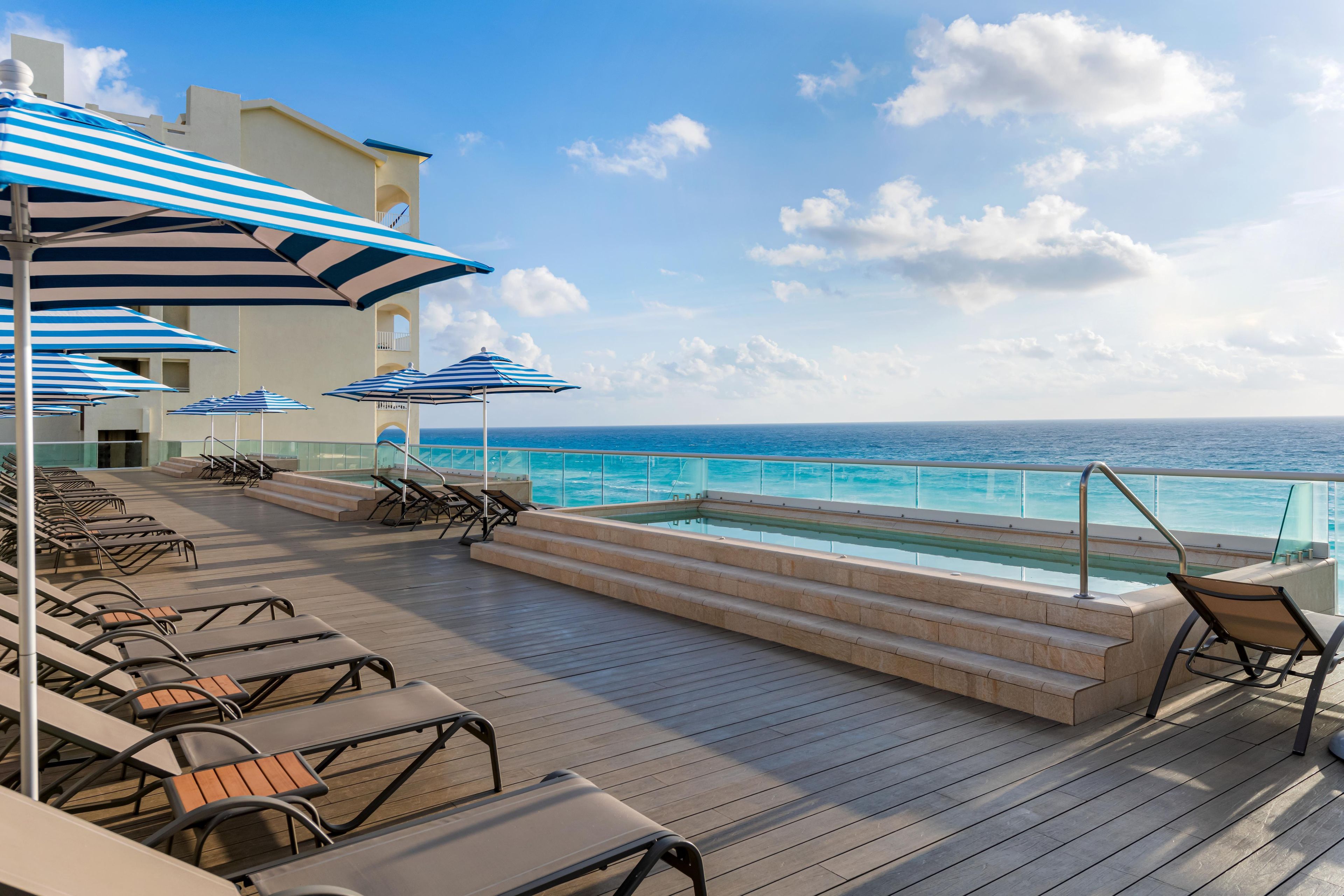 A deck with lounge chairs and umbrellas overlooking a pool and the ocean.