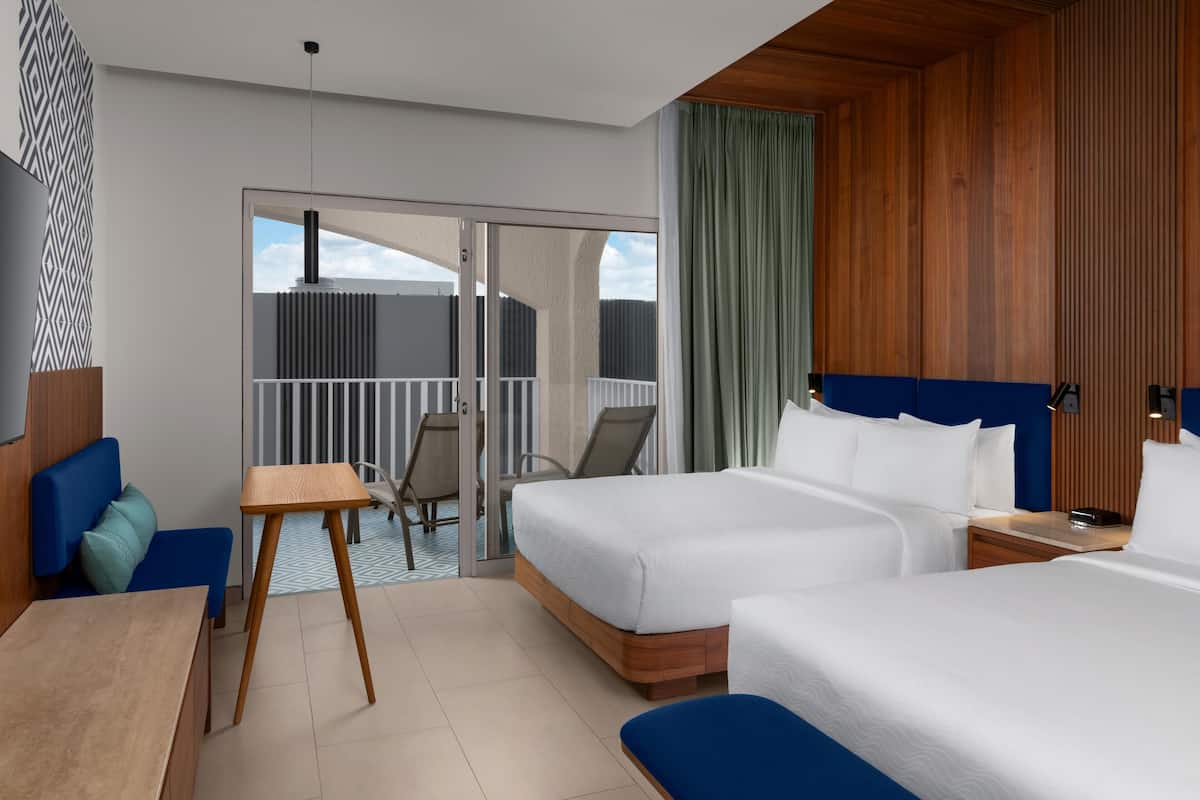 A hotel room with two beds, a desk, a chair, a small table, and a balcony with a view.