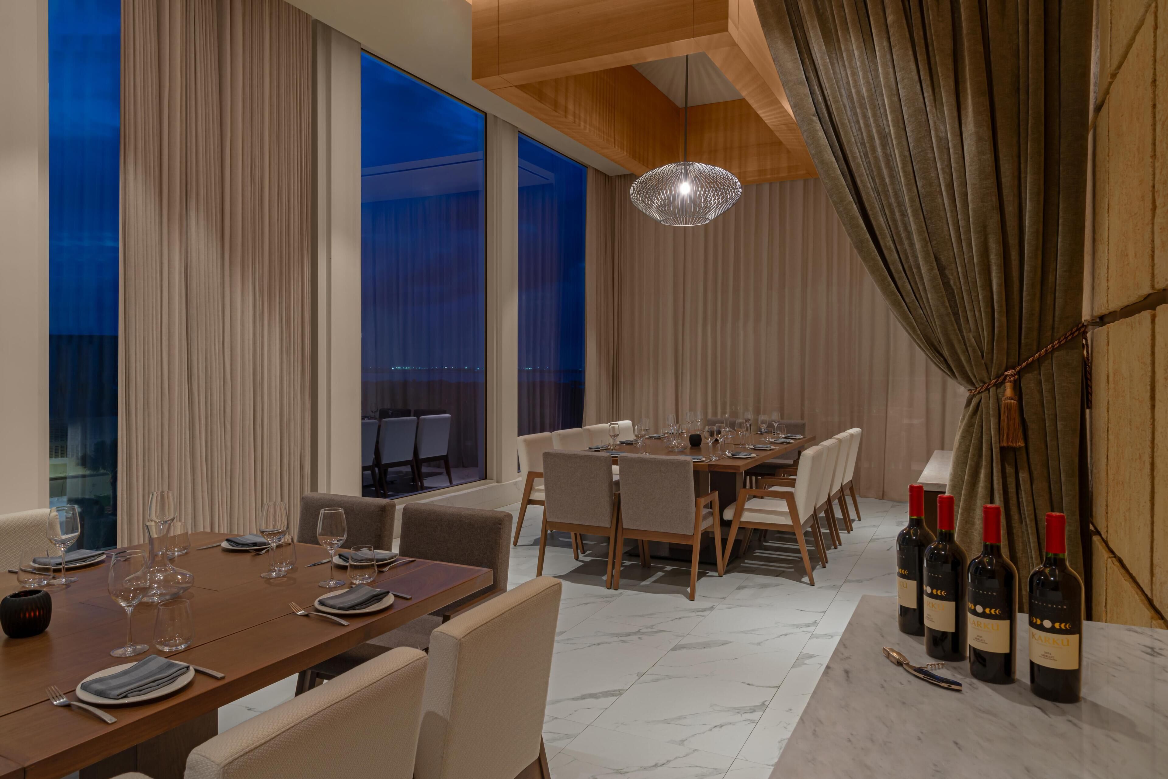 A modern dining area with a long table set for a meal, large windows, and a view of the cityscape.