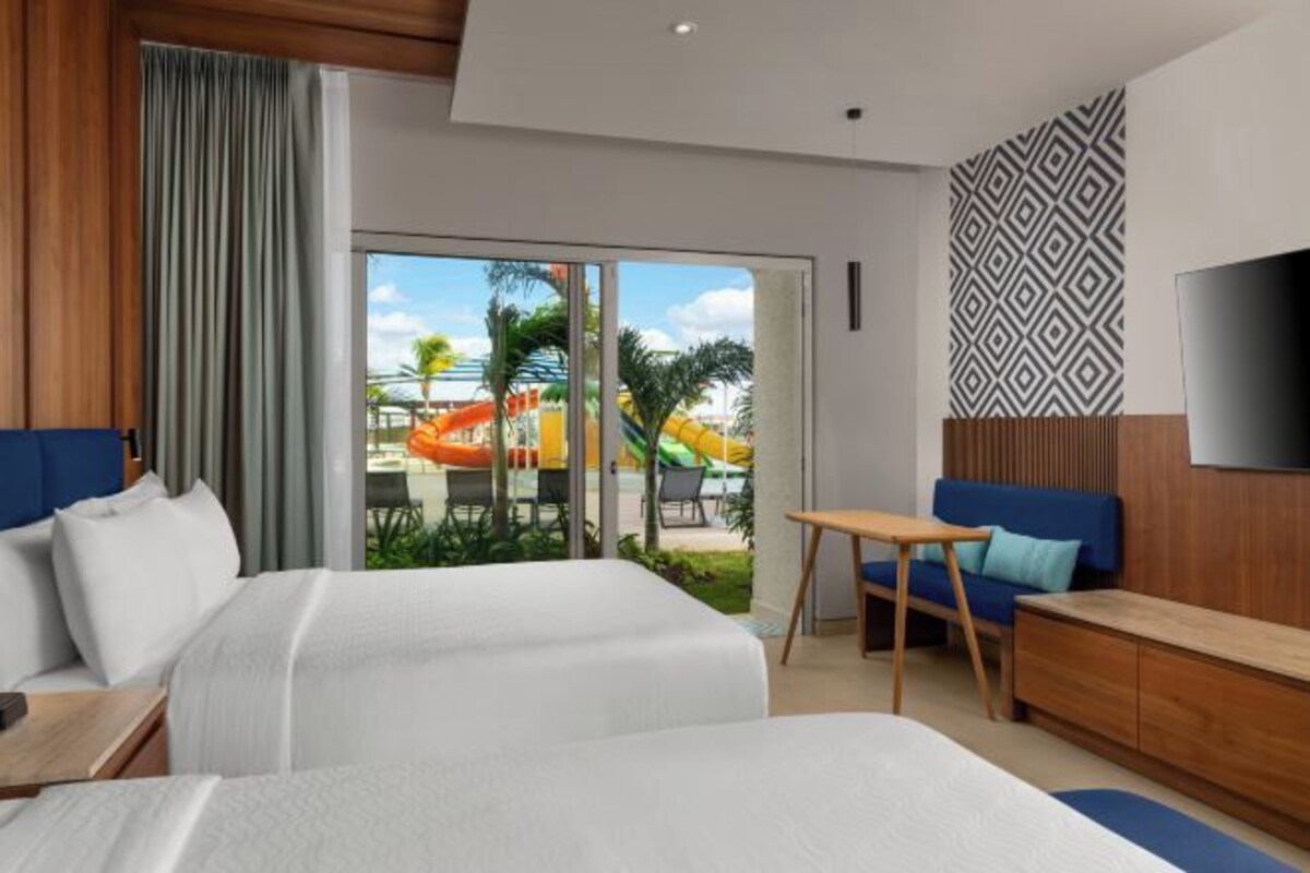 A hotel room with a bed, a desk, a TV, and a view of a pool area.