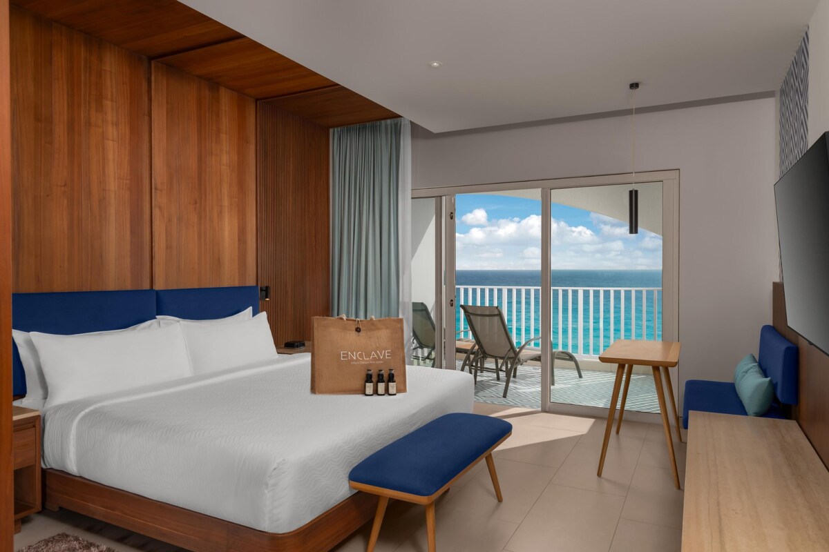 A hotel room with a bed, a bench, a table, and a view of the ocean.