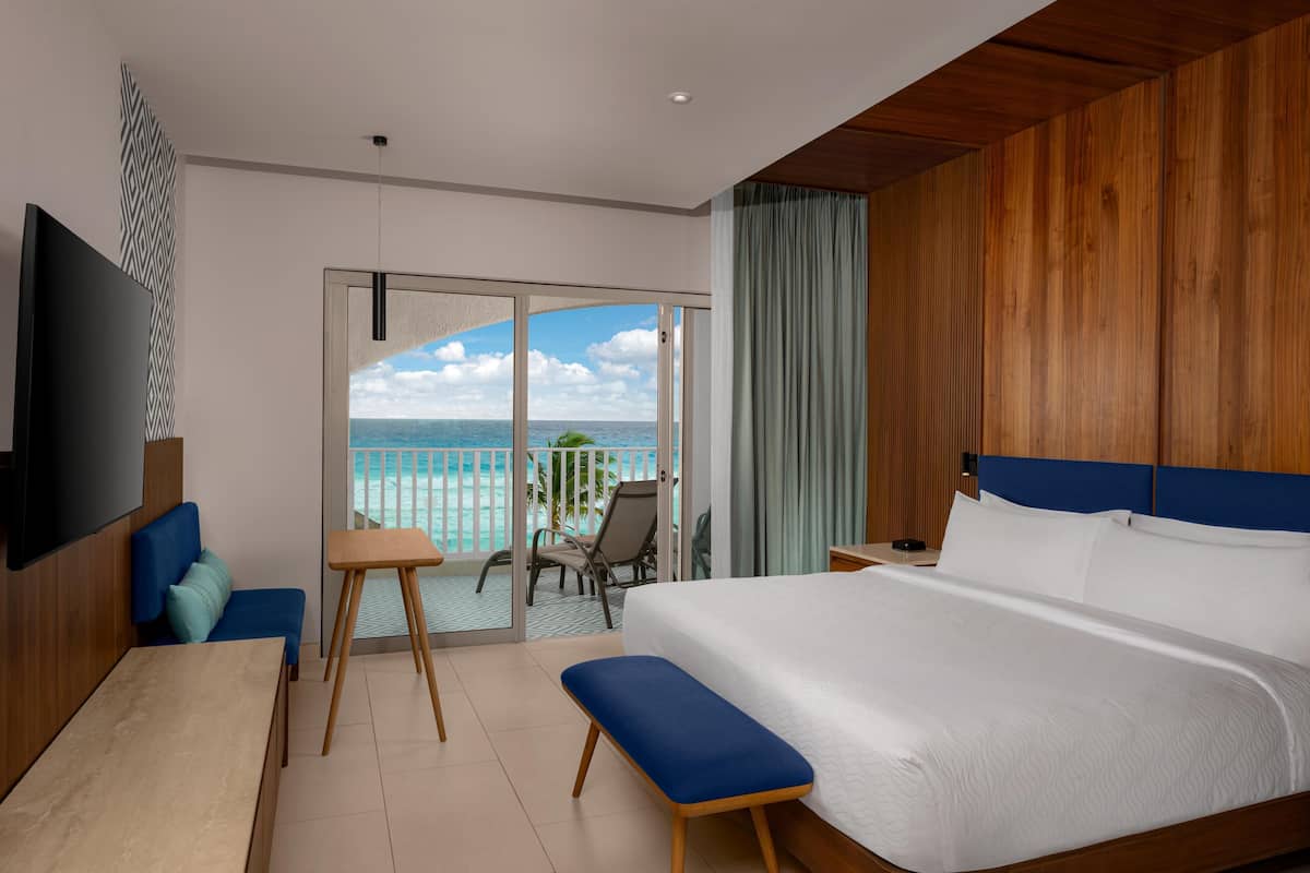 A modern hotel room with a large bed, a desk, a chair, and a balcony with a view of the ocean.