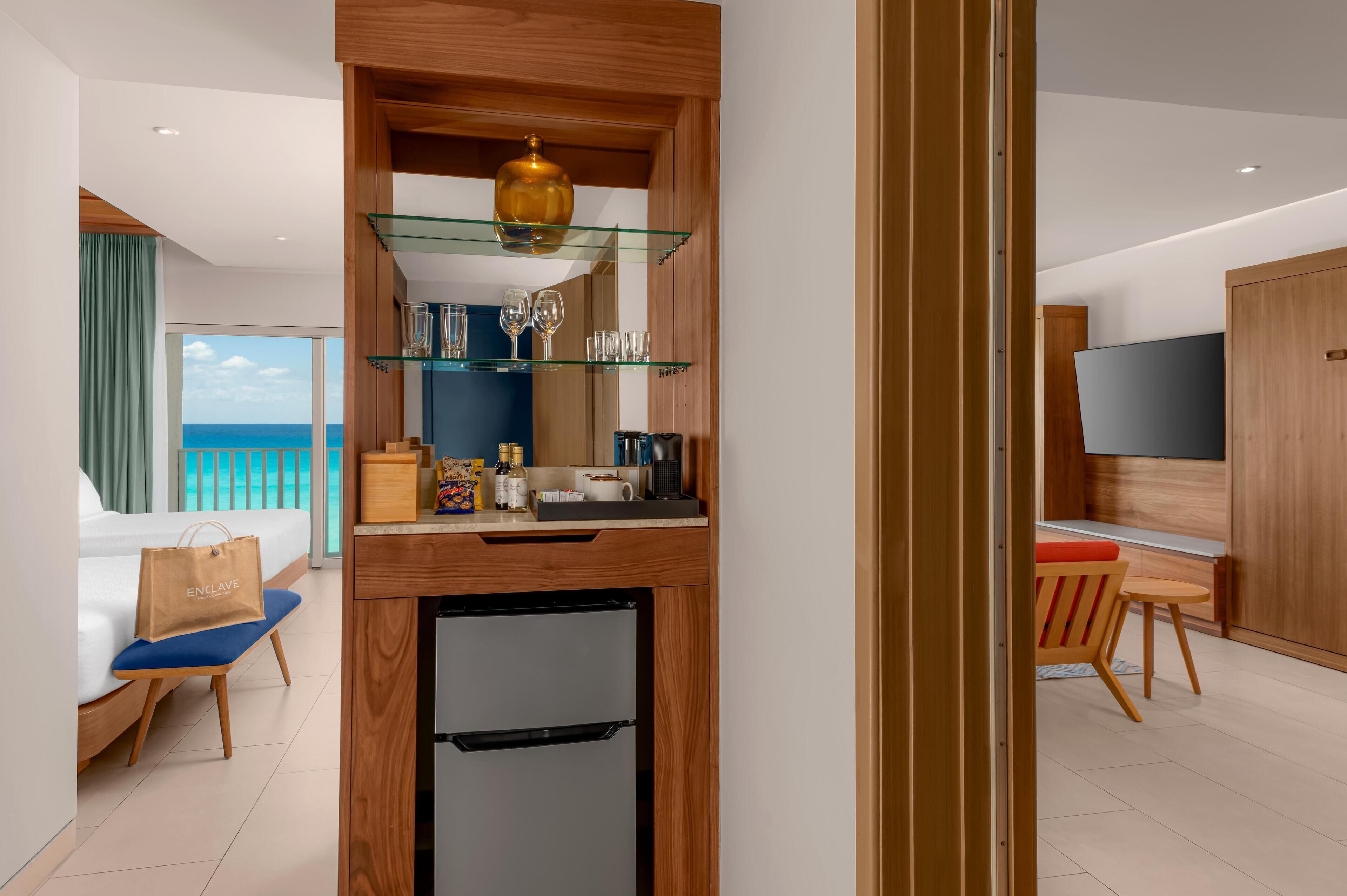 Une chambre d’hôtel comprenant un lit, un petit bureau, un mini-réfrigérateur, une étagère en verre avec des objets décoratifs, une télévision et une vue sur l’océan.