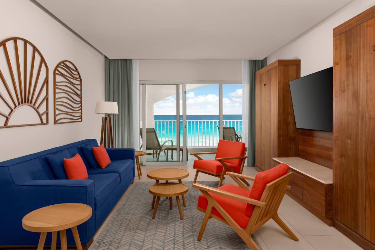 A hotel room with a blue sofa, red chairs, a wooden TV stand, and a view of the ocean through large windows.
