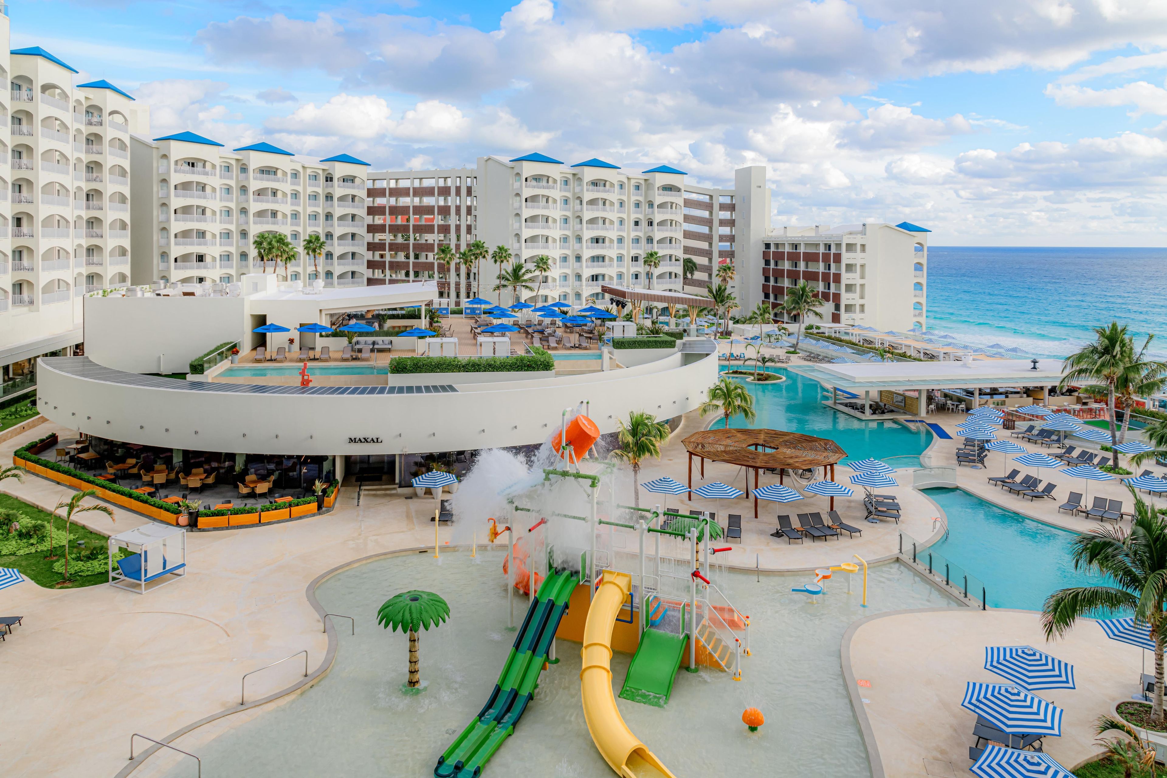 A resort with multiple swimming pools, a water slide, and a beach area with lounge chairs and umbrellas.
