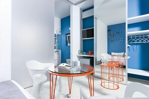 Una habitación de hotel moderna con una pared azul de acento, una mesa de vidrio con patas naranjas, sillas blancas y un sofá blanco.