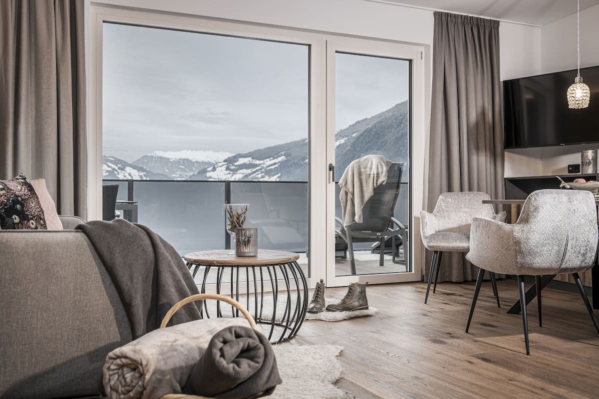 Un salon moderne comprenant un canapé, des fauteuils, une petite table ronde et un téléviseur. Une grande porte-fenêtre coulissante donne accès à un balcon offrant une vue sur les montagnes.