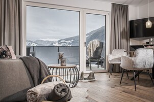 A modern living room with a sofa, chairs, a small round table, and a TV. A large sliding glass door leads to a balcony with a mountain view.