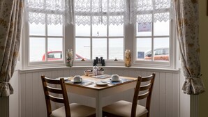 A corner dining area with a table set for two, two chairs, and a view of the sea through large windows with lace curtains. - The Seaspray (Weymouth)