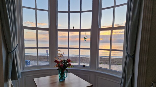 A room with a bay window overlooking a beach. There is a table with a vase of flowers and two chairs. - The Seaspray (Weymouth)