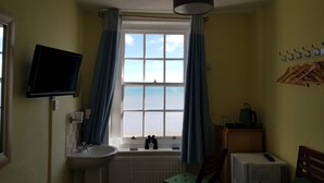 A small room with a window overlooking the sea, a sink, a chair, a table with a box, and a television mounted on the wall. - The Seaspray (Weymouth)