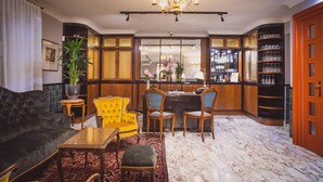 Un vestíbulo de hotel con un sofá oscuro, un sillón amarillo y dos sillas de madera. Se observa un área de bar con estantes llenos de vasos y botellas al fondo.