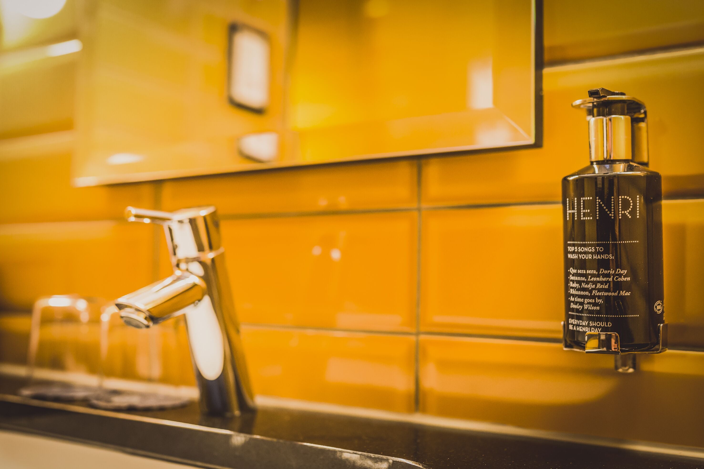 A bathroom with a yellow tiled wall, a chrome faucet, and a bottle of Henri hand soap.