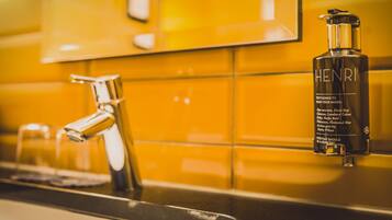 A bathroom with a yellow tiled wall, a chrome faucet, and a bottle of Henri hand soap.