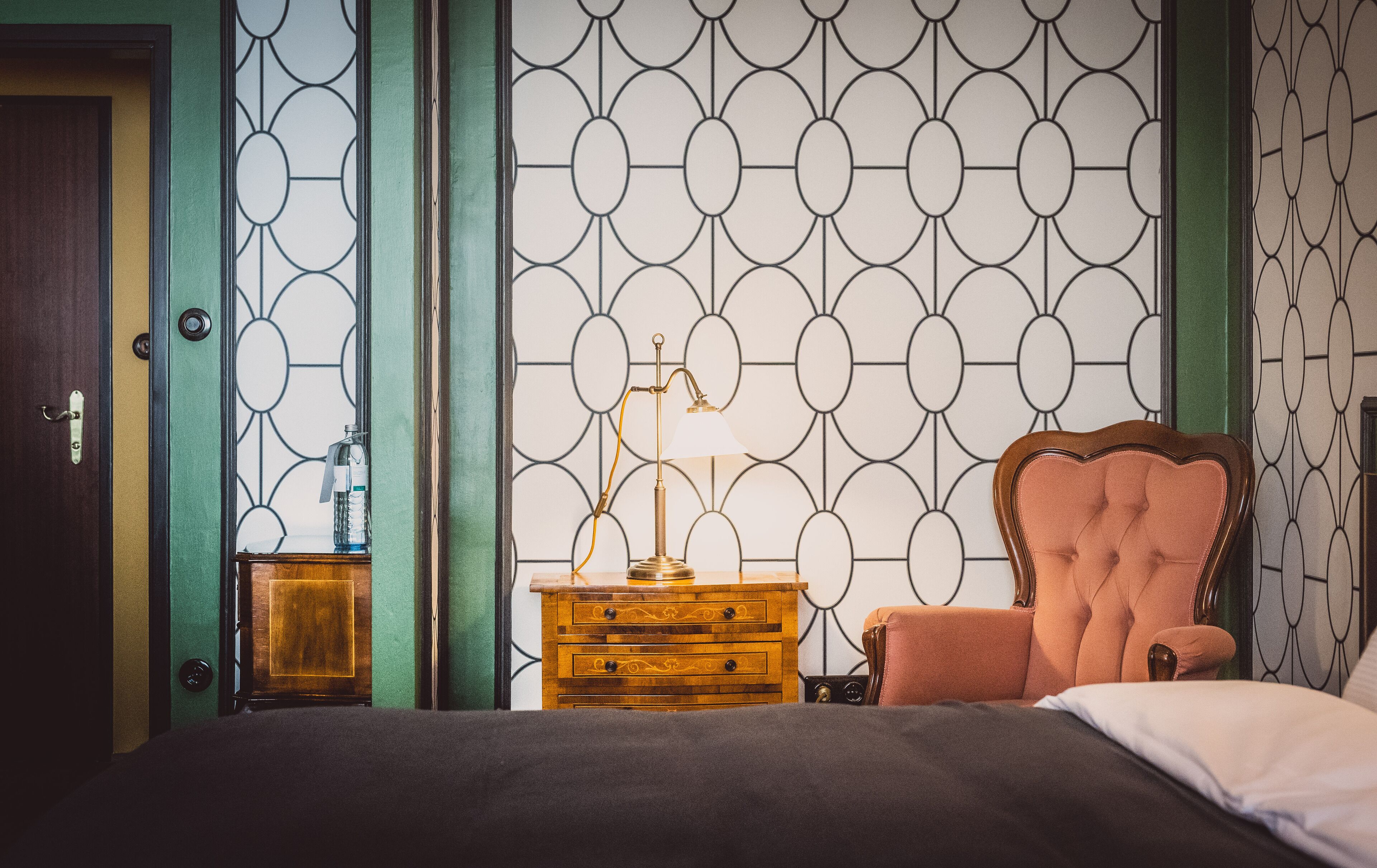 Un dormitorio con una cama, una mesita de noche de madera, una lámpara, una silla y un papel tapiz estampado.