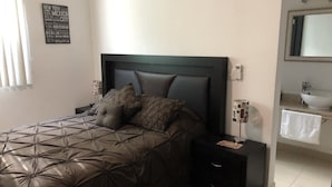 A bedroom with a bed, a nightstand, a bedside table, a mirror, and a wall-mounted sign listing cities.