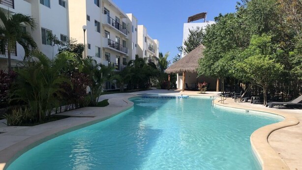 A swimming pool with a curved edge, surrounded by palm trees and a building with balconies.