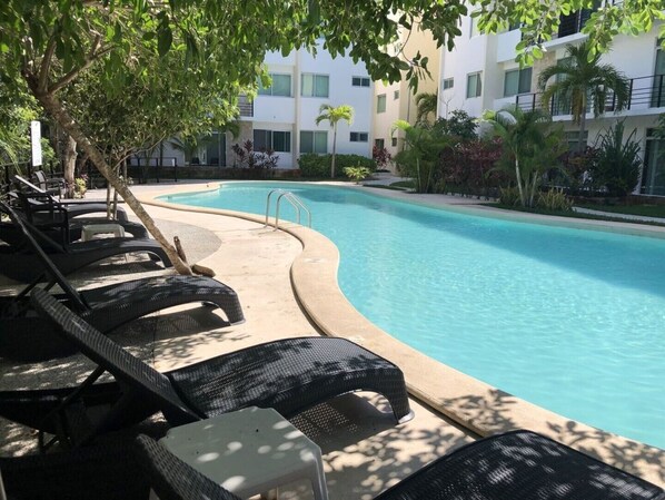A swimming pool with lounge chairs and a small table, surrounded by greenery and a building in the background. - PLAYA SUITES I POOL VIEW (Carmen)