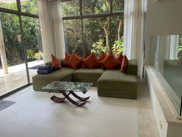 Un salon moderne avec un canapé d’angle, une table basse en verre et de grandes fenêtres donnant sur un paysage verdoyant.