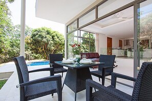 An outdoor dining area with wicker furniture and a glass table, overlooking a swimming pool and lush greenery.