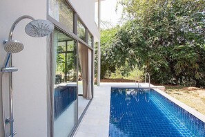 A modern outdoor pool area with a glass-enclosed shower, surrounded by lush greenery.