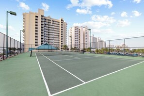 Uma quadra de tênis ao ar livre com rede, cercada por edifícios residenciais altos e um céu claro.