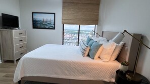 Relax in this serene bedroom with a large bed, nightstand, and TV, offering a stunning city and beach view.