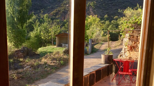 Ventana de madera con vistas a un paisaje rural, que incluye un patio de piedra, una silla roja y una mesa pequeña.