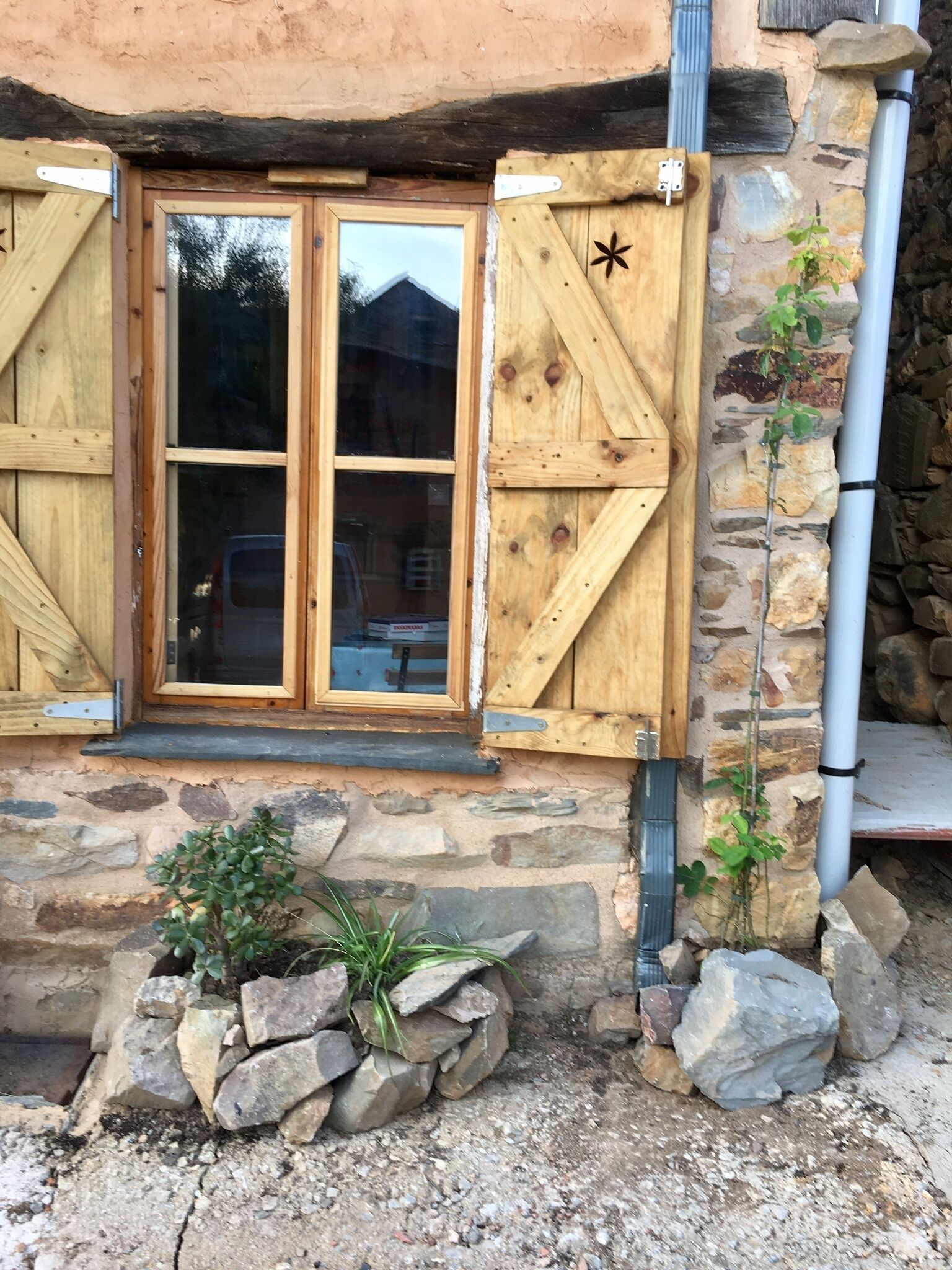 A rustic stone building with wooden shutters, a window with a star-shaped cutout, and a small potted plant beside the entrance.