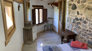 Una habitación pequeña y rústica con pared de piedra, vigas de madera en el techo, un escritorio, una silla y una cama con un edredón enrollado.