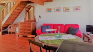 Un salon chaleureux avec un escalier en bois, un canapé rouge, une table ronde recouverte d’une nappe verte et un téléviseur posé sur un meuble.