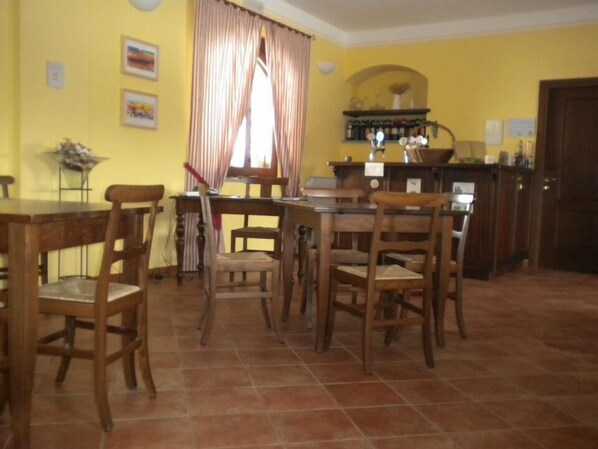 A dining area with a wooden table and chairs, a window with curtains, and a kitchen area with a counter and cabinets. - B&B Cascina Orto (Pasturana)