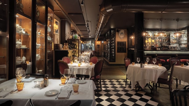A restaurant with checkered flooring, tables set with white tablecloths, and shelves with glassware. - Speronari Suites (Milan)