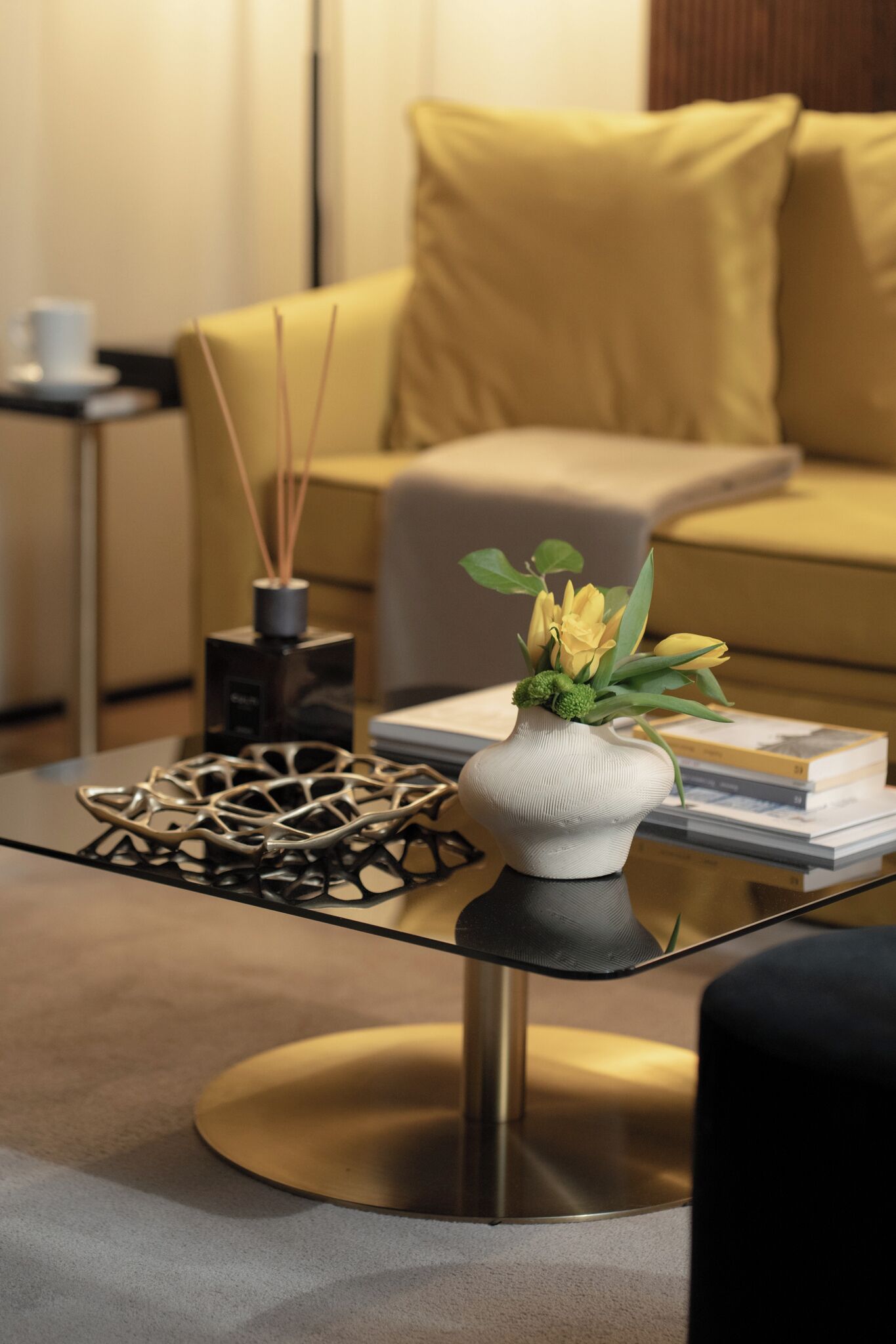 Una sala de estar moderna con una mesa de centro de vidrio, un jarrón con flores, una escultura metálica decorativa y un sofá con cojines.