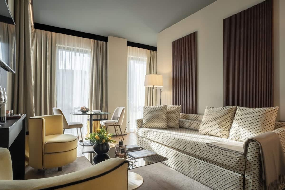 A modern hotel room with a sofa, armchair, dining table, and lamp.