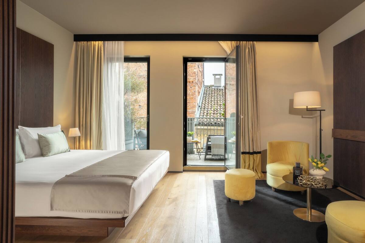 A hotel room with a bed, a sofa, a chair, a small table, and a balcony with a view of buildings.