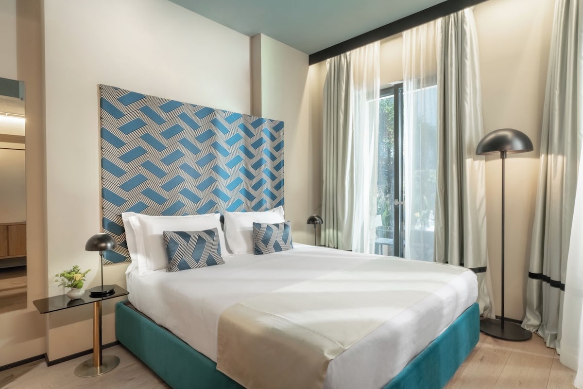 A hotel room with a bed, a nightstand, a lamp, and a window with curtains.