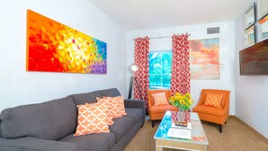 Ein Wohnzimmer mit grauem Sofa, orangefarbenen Sesseln, einem Couchtisch und einem farbenfrohen abstrakten Gemälde.