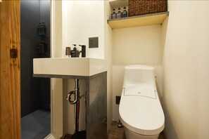 A bathroom with a white sink, toilet, and shower area. There are shelves with toiletries and a basket on the wall. - mizuka Imaizumi 4 - unmanned hotel - (Fukuoka)