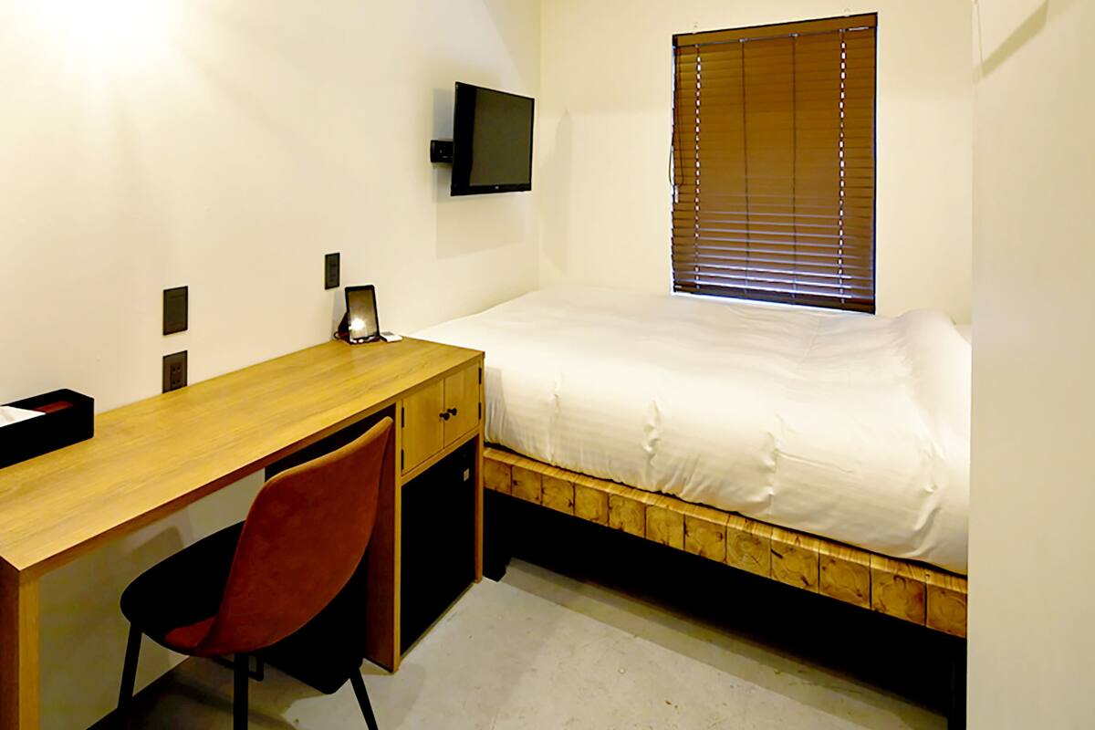 A bedroom with a wooden bed, a desk, a chair, a television on the wall, and a window with blinds.