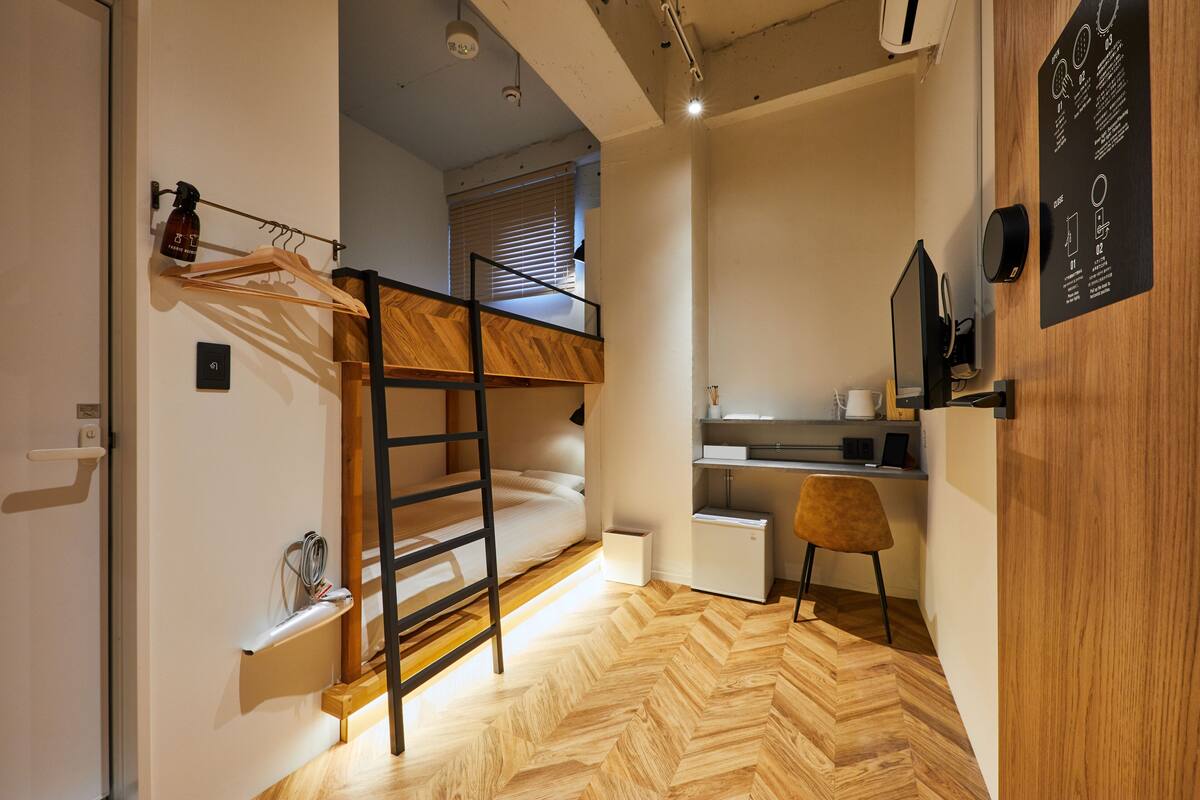Une pièce compacte comprenant un lit superposé, un bureau et une chaise, avec un parquet en bois disposé en chevrons et un panneau mural en bois.